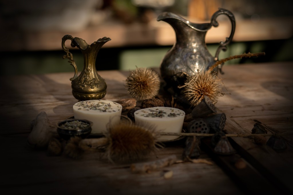 En bild på två vita, runda tvålar med torkade lavendelblommor på som ligger på ett träbord med sirliga kannor och torkade blommor omkring sig.