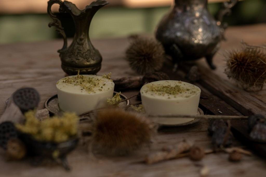 En bild på två vita, runda tvålar med torkad älgört på som ligger på ett träbord med sirliga kannor och torkade blommor omkring sig.