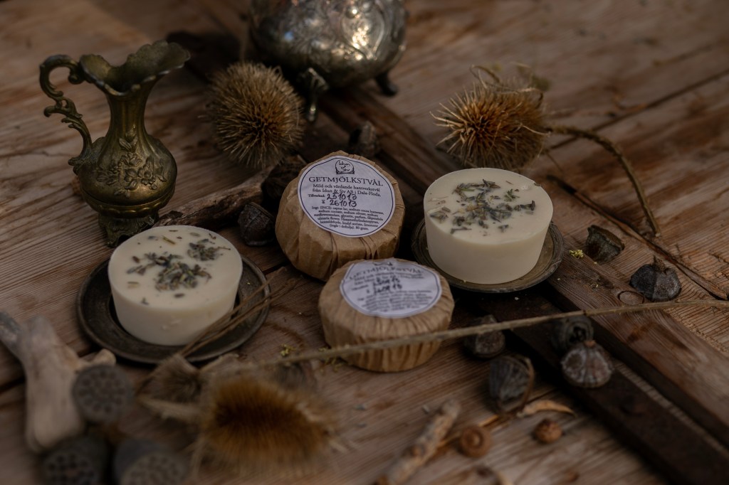 En bild på två vita, runda tvålar med torkade lavendelblommor på och två tvålar inslagna i brunt papper med en vit etikett. De ligger på ett träbord med sirliga kannor och torkade blommor omkring sig.