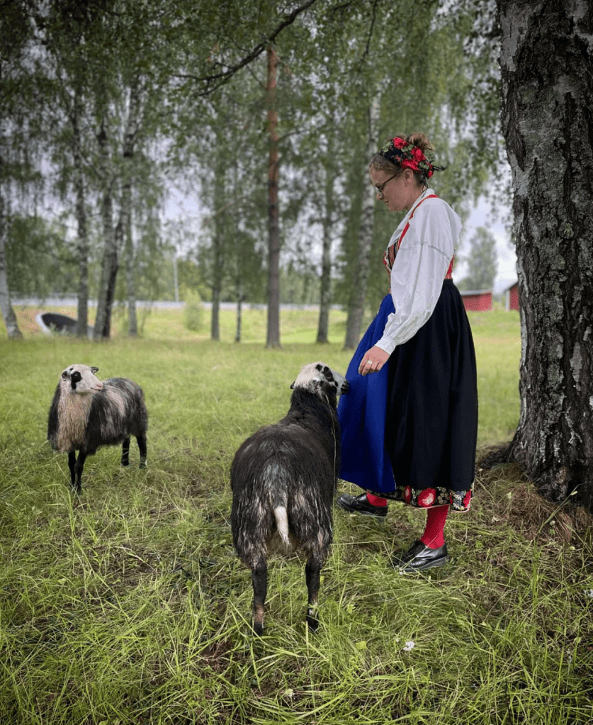 En bild på en kvinna iklädd folkdräkt från Dala-Floda vid en björk i en hage med två gulliga lamm.